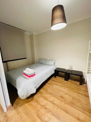 Una habitación pequeña con cama individual, piso de madera, colcha gris, mesita de noche y una lámpara colgante.