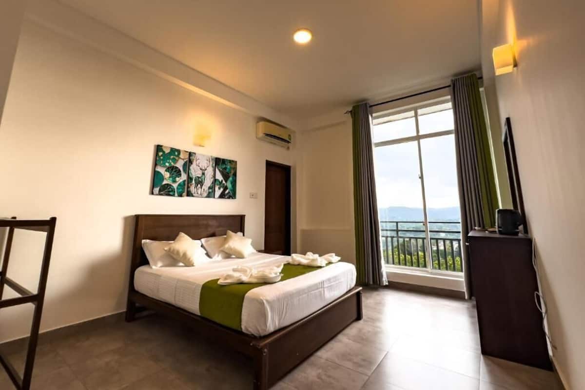 Habitación de hotel con cama, una silla, un cuadro, equipo de aire acondicionado, ventana con cortinas y balcón con vistas.