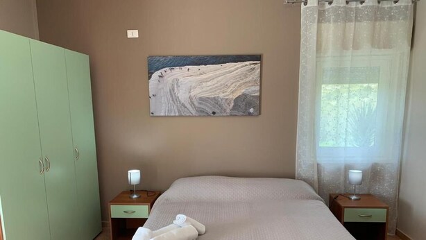 Une chambre à coucher avec un lit, deux tables de chevet, une armoire verte, un tableau au mur et une fenêtre avec des rideaux.