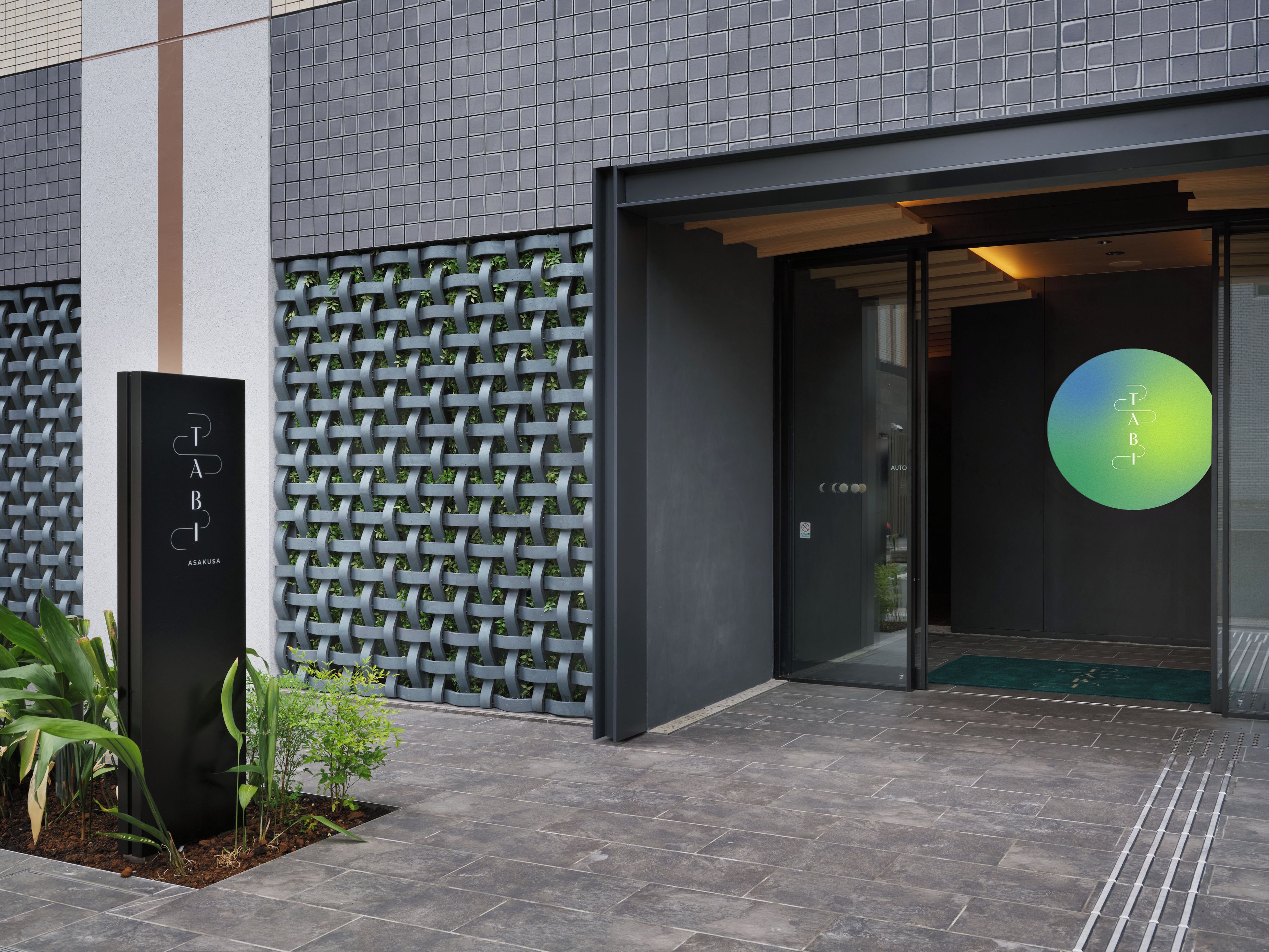 A modern building entrance with a metal lattice wall, a black sign with 'TABI ASAKUSA', and a green circular logo on the door.