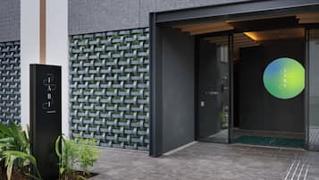 Una entrada moderna de edificio con un muro de celosía metálica, un letrero negro que dice 'TABI ASAKUSA' y un logotipo circular verde en la puerta.