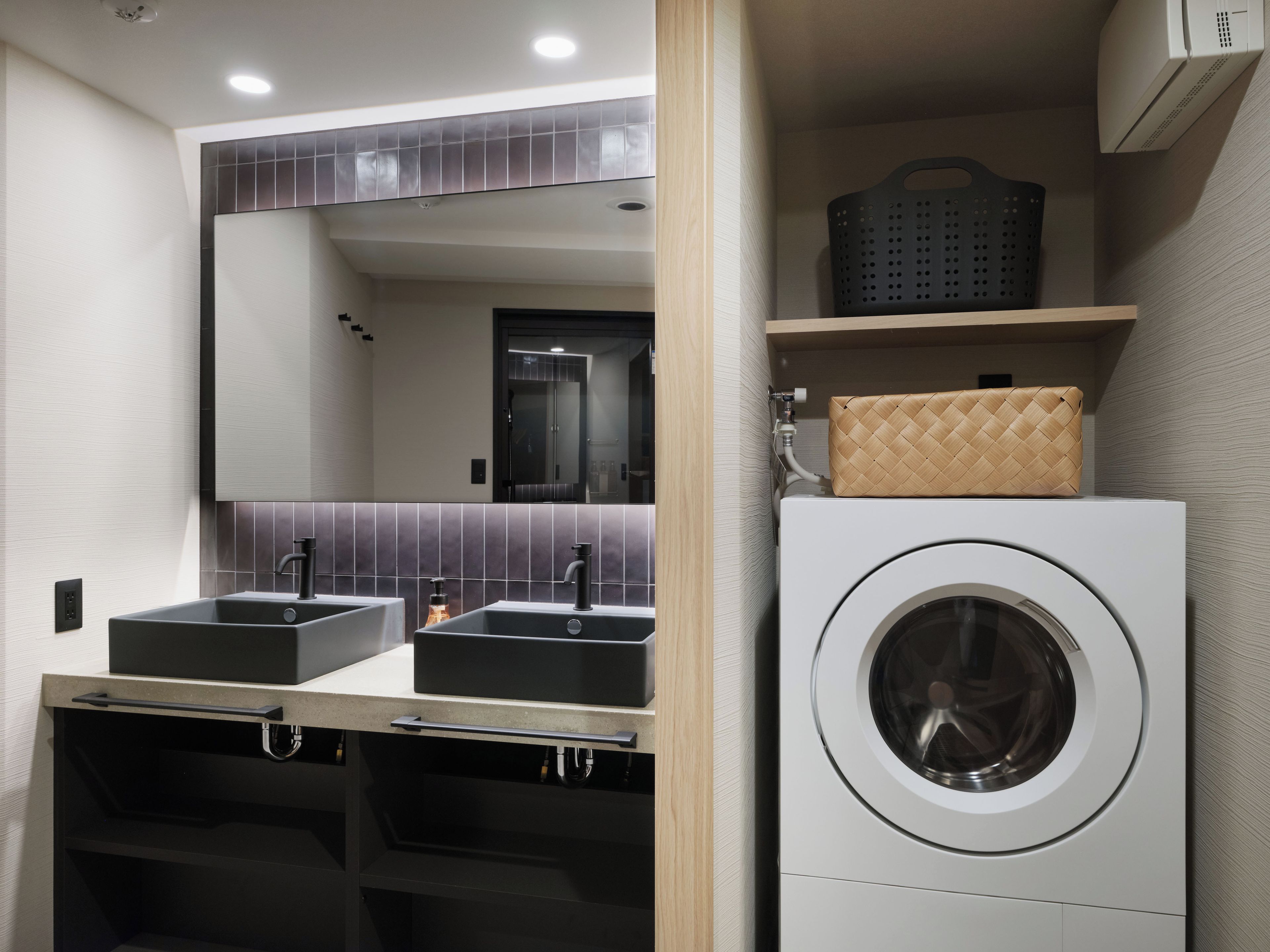 A modern bathroom with dual sinks, a large mirror, a washing machine, and a basket on a shelf.