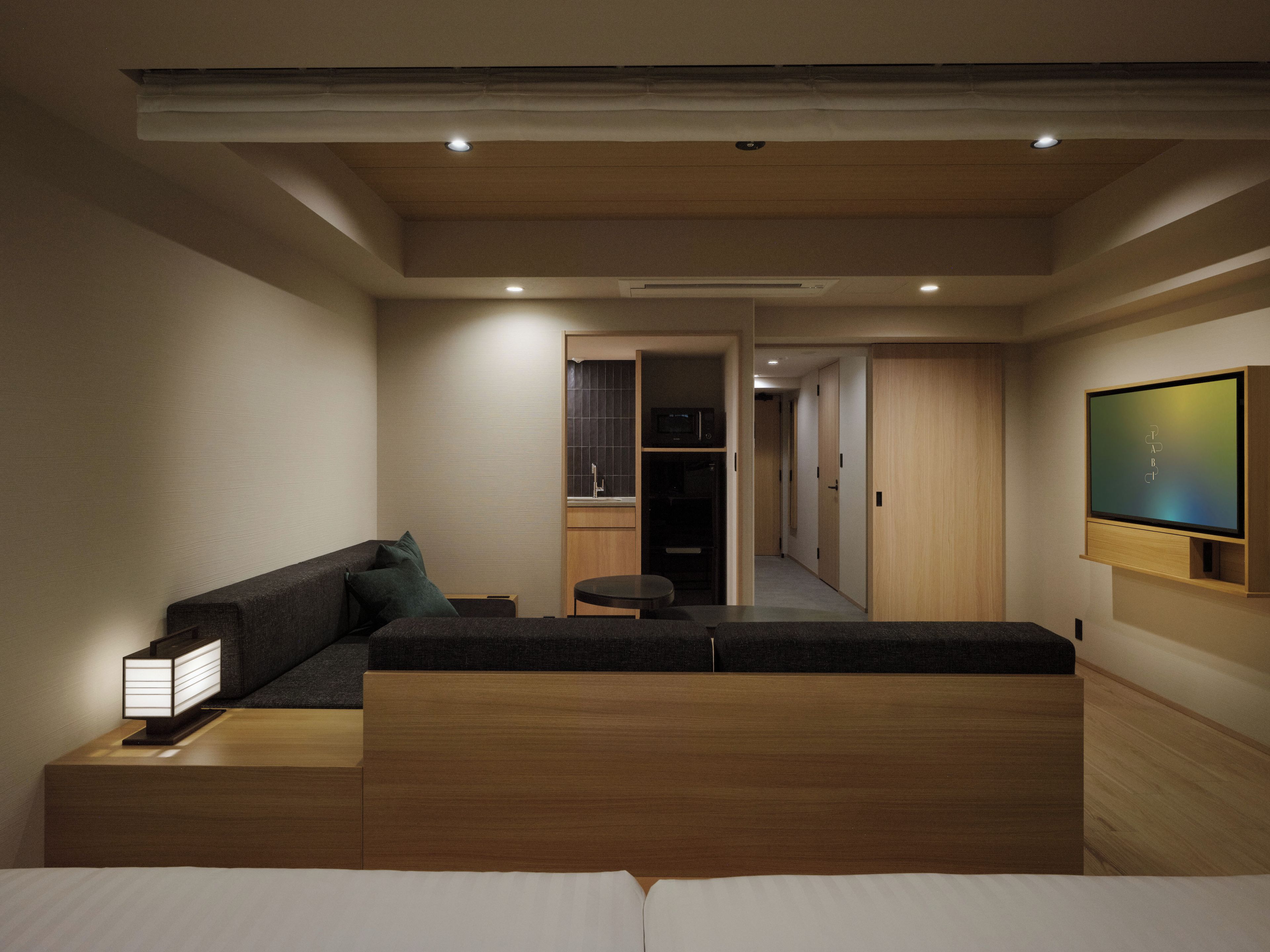 A modern hotel room with a sofa, a small table, a TV mounted on the wall, and a hallway leading to a bathroom.