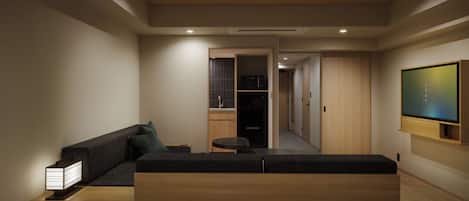 Una habitación de hotel moderna con sofá, una mesita, televisor montado en la pared y un pasillo que conduce a un baño.