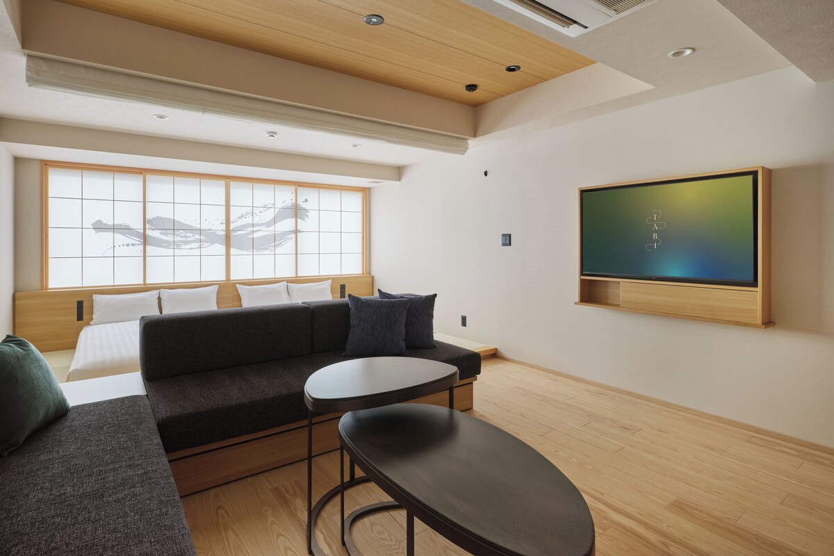 A modern hotel room with a sofa, a small table, a TV mounted on the wall, and a large window with a view.