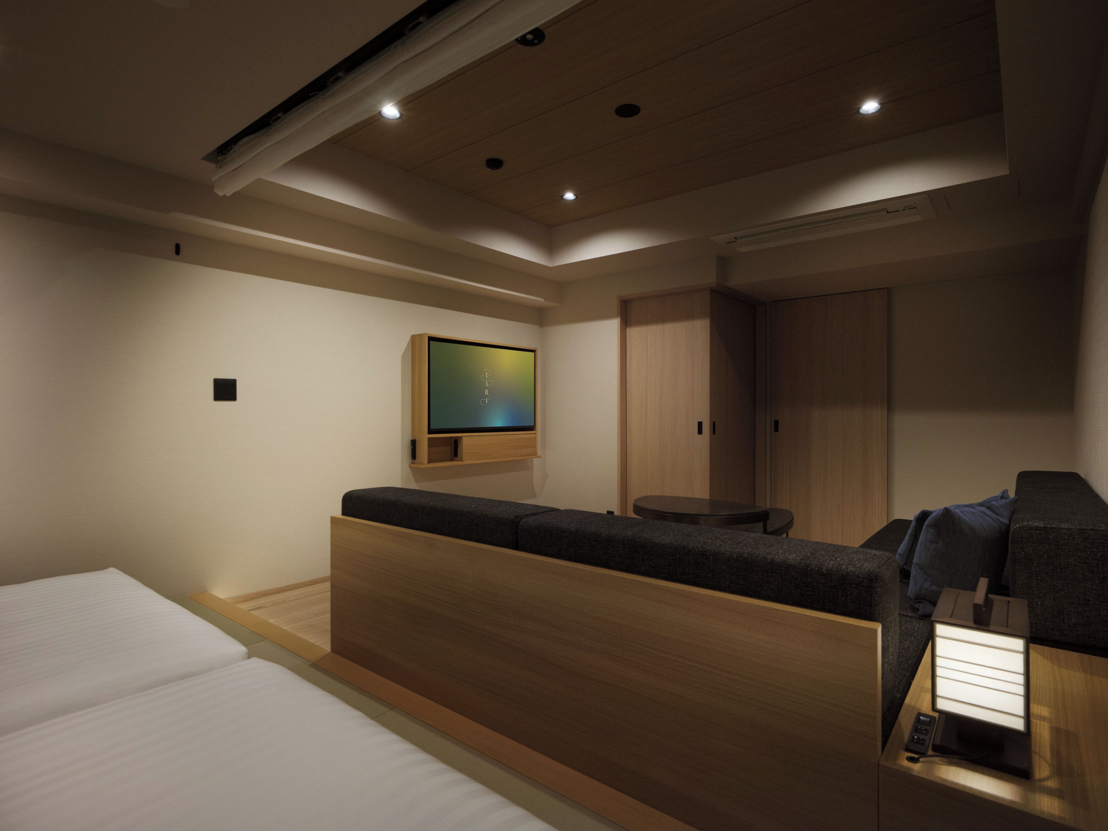 A modern hotel room with a bed, a seating area, a TV mounted on the wall, and a wardrobe.