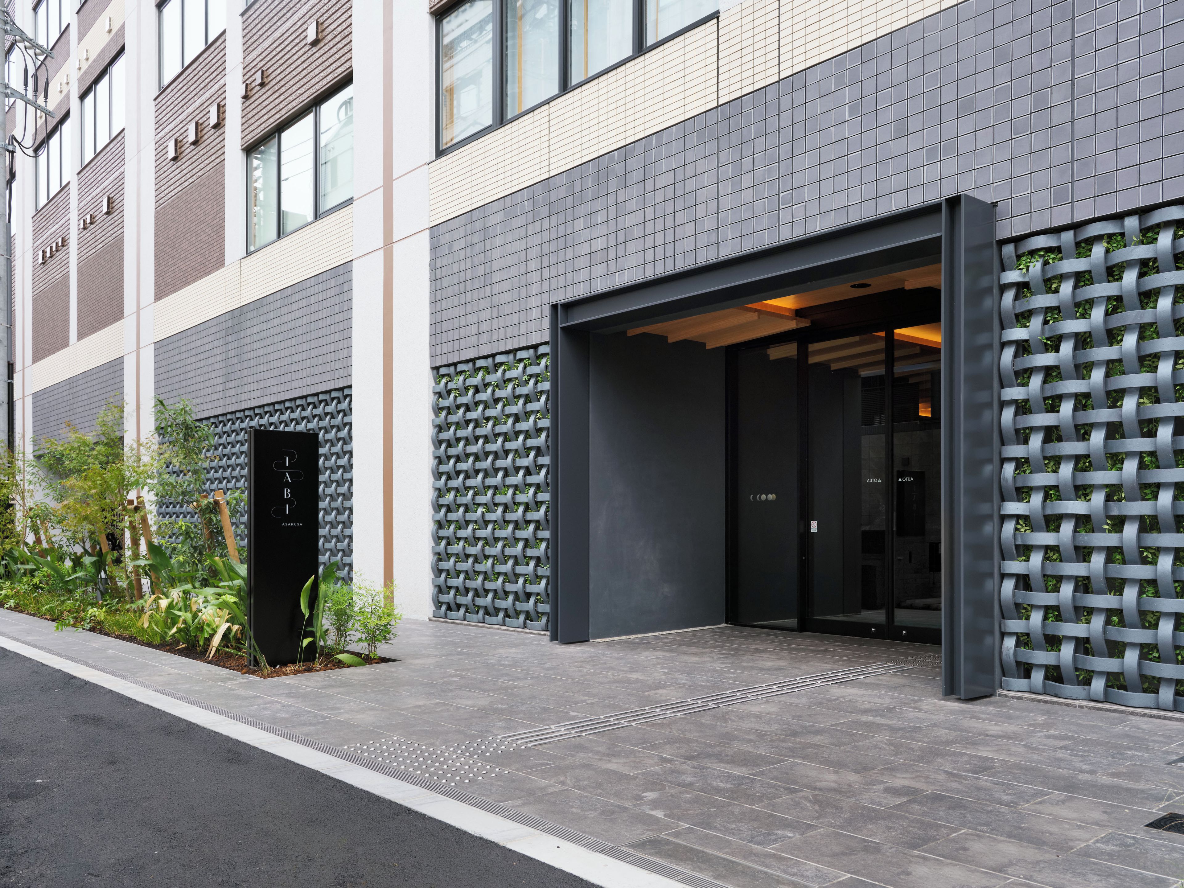 A modern building entrance with a dark facade, a metal gate, and a small garden area with plants.
