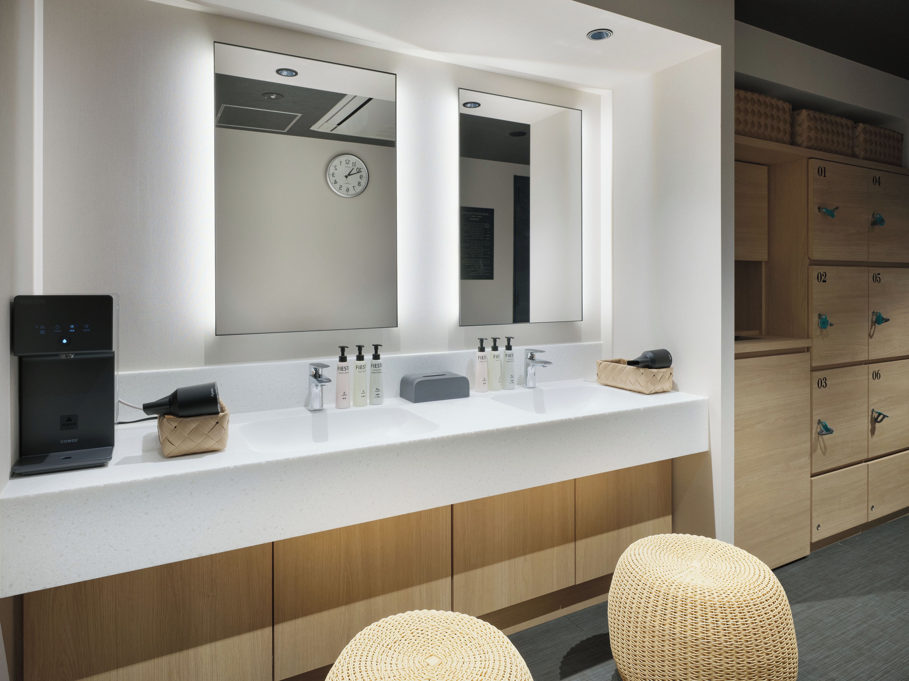 A modern bathroom with a white countertop, two sinks, a mirror, a clock, and a coffee machine.
