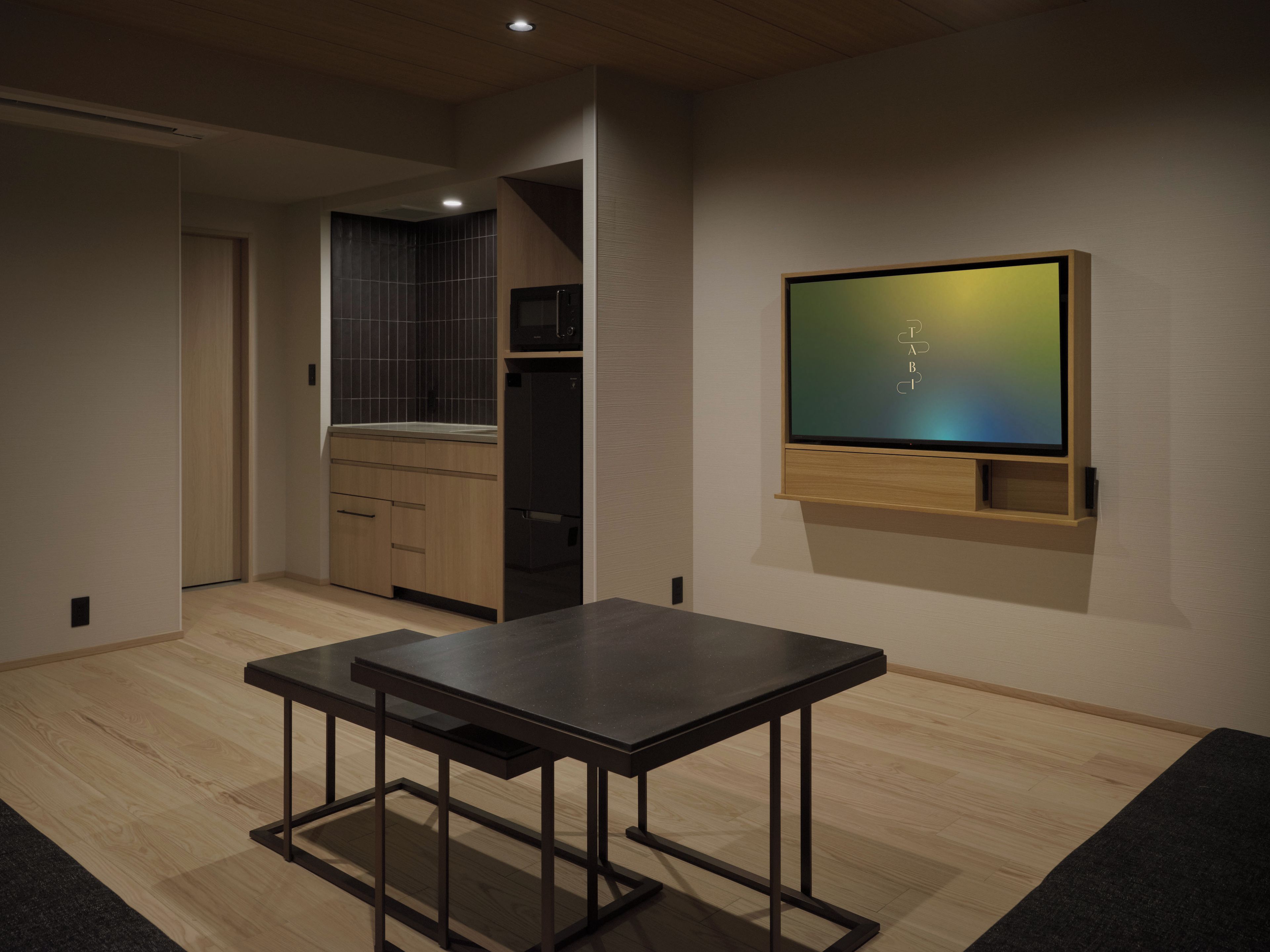 Una sala moderna con un televisor de pantalla plana montado en la pared, una mesa de madera oscura y una cocina con armarios oscuros.