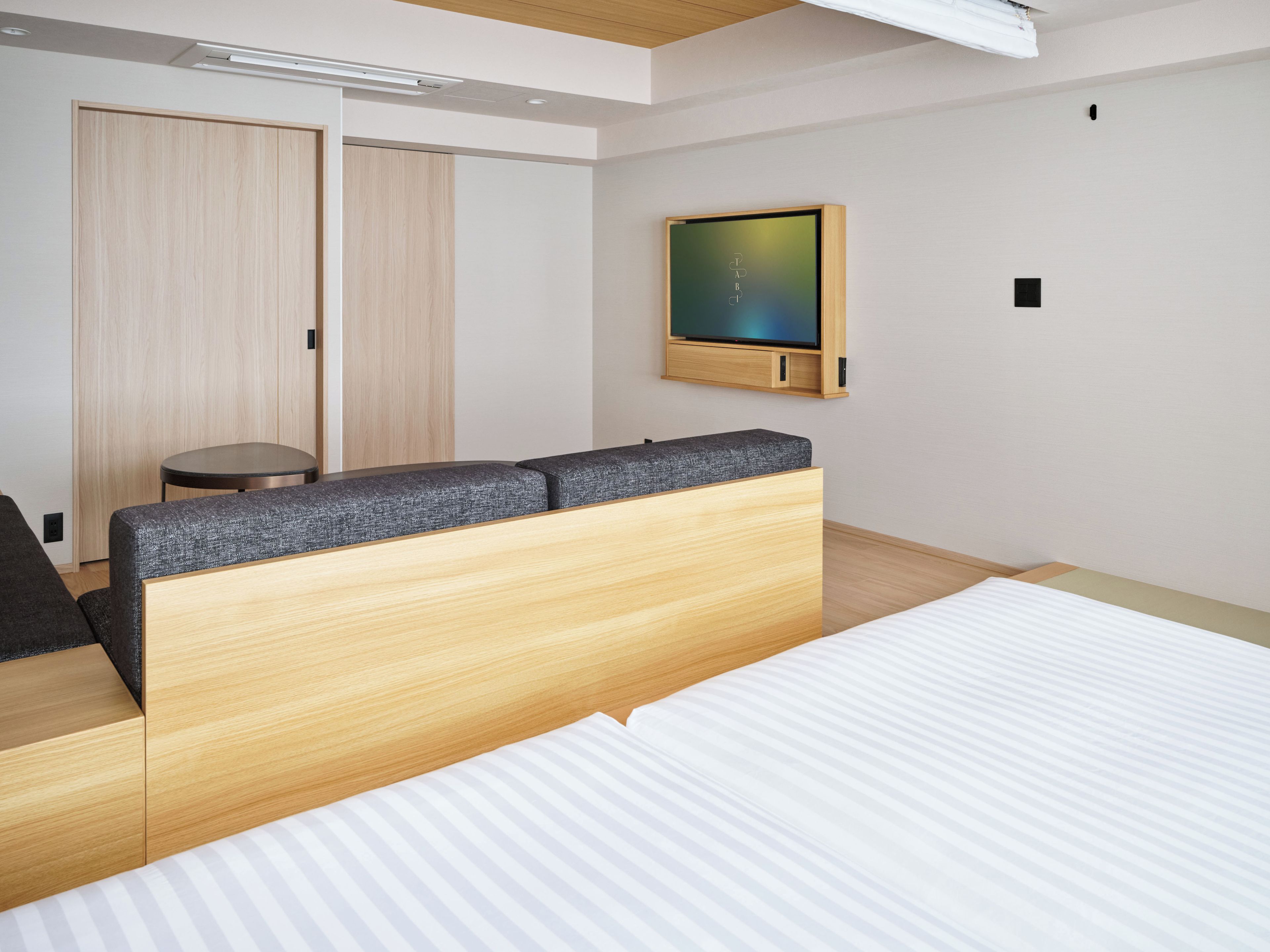 A modern hotel room with a large bed, a wooden headboard, a flat-screen TV mounted on the wall, and a small round table.