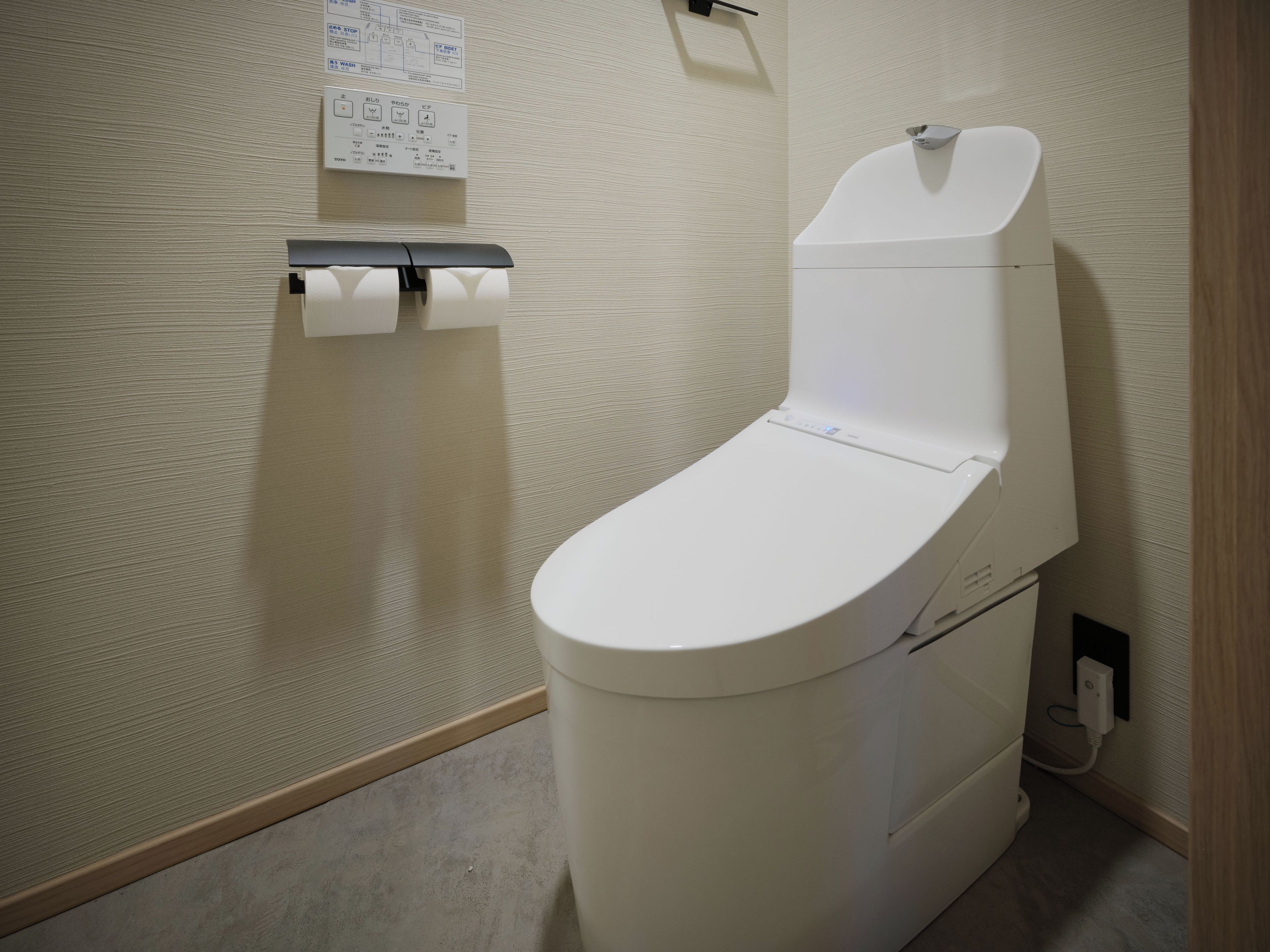 Un inodoro moderno con panel de control, dispensador de papel higiénico y botón de descarga en la pared.