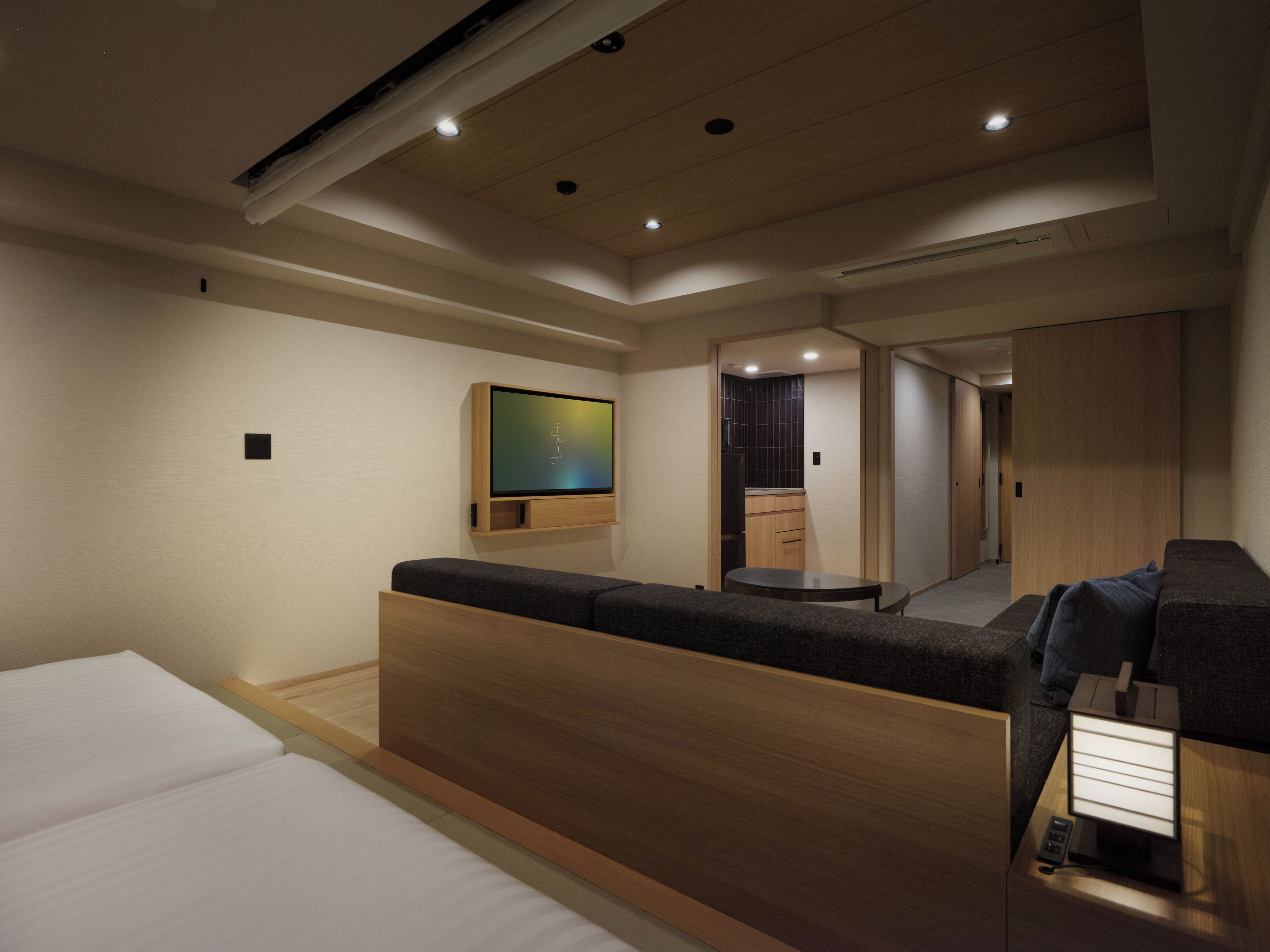 A modern hotel room with a bed, a seating area, a television, and a kitchenette.