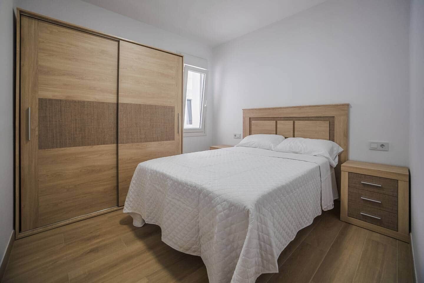 A bedroom with a bed, a wooden wardrobe, a bedside table, and a window.