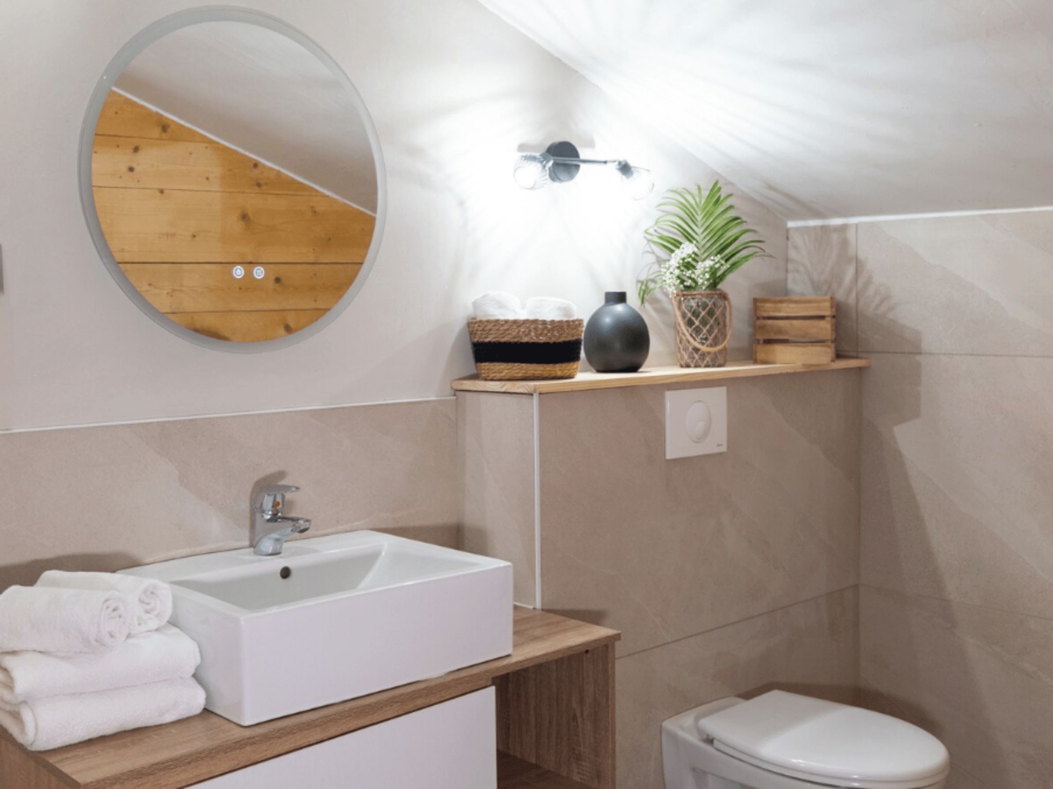 Ein modernes Badezimmer mit einem weißen Waschbecken, einem runden Spiegel, einer Toilette und einem Regal mit Pflanze und Korb.