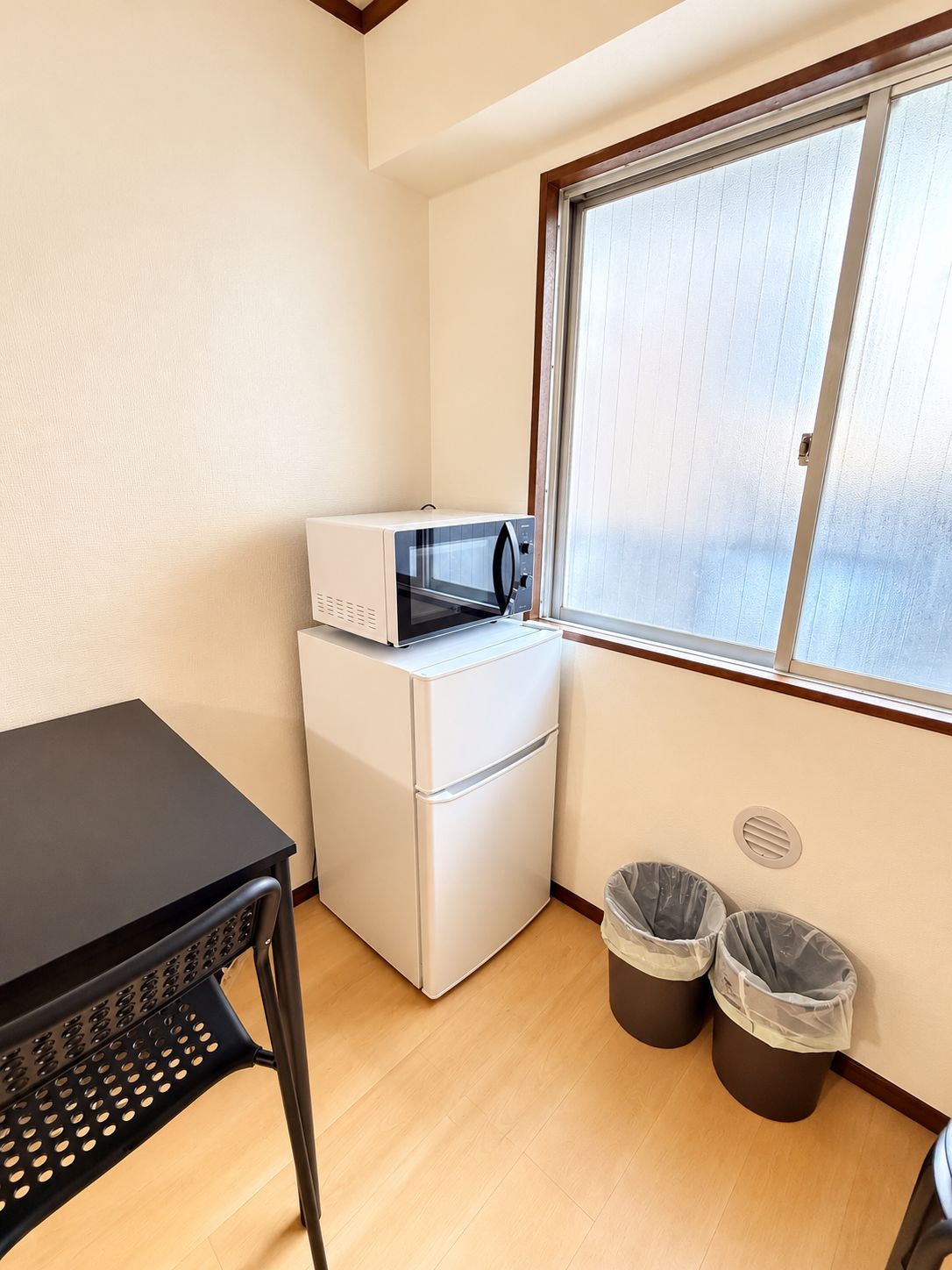 A compact kitchenette with a microwave, refrigerator, and a small table. There are two trash bins and a window providing natural light.
