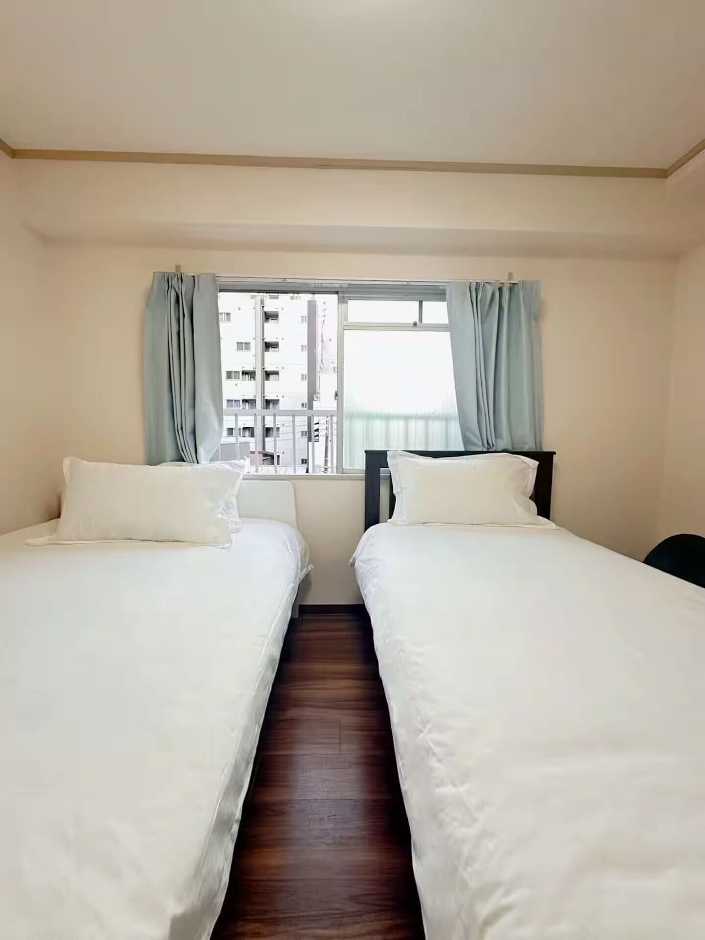 Une chambre d’hôtel avec deux lits, une fenêtre avec des rideaux, une vue sur des immeubles et un plancher en bois.