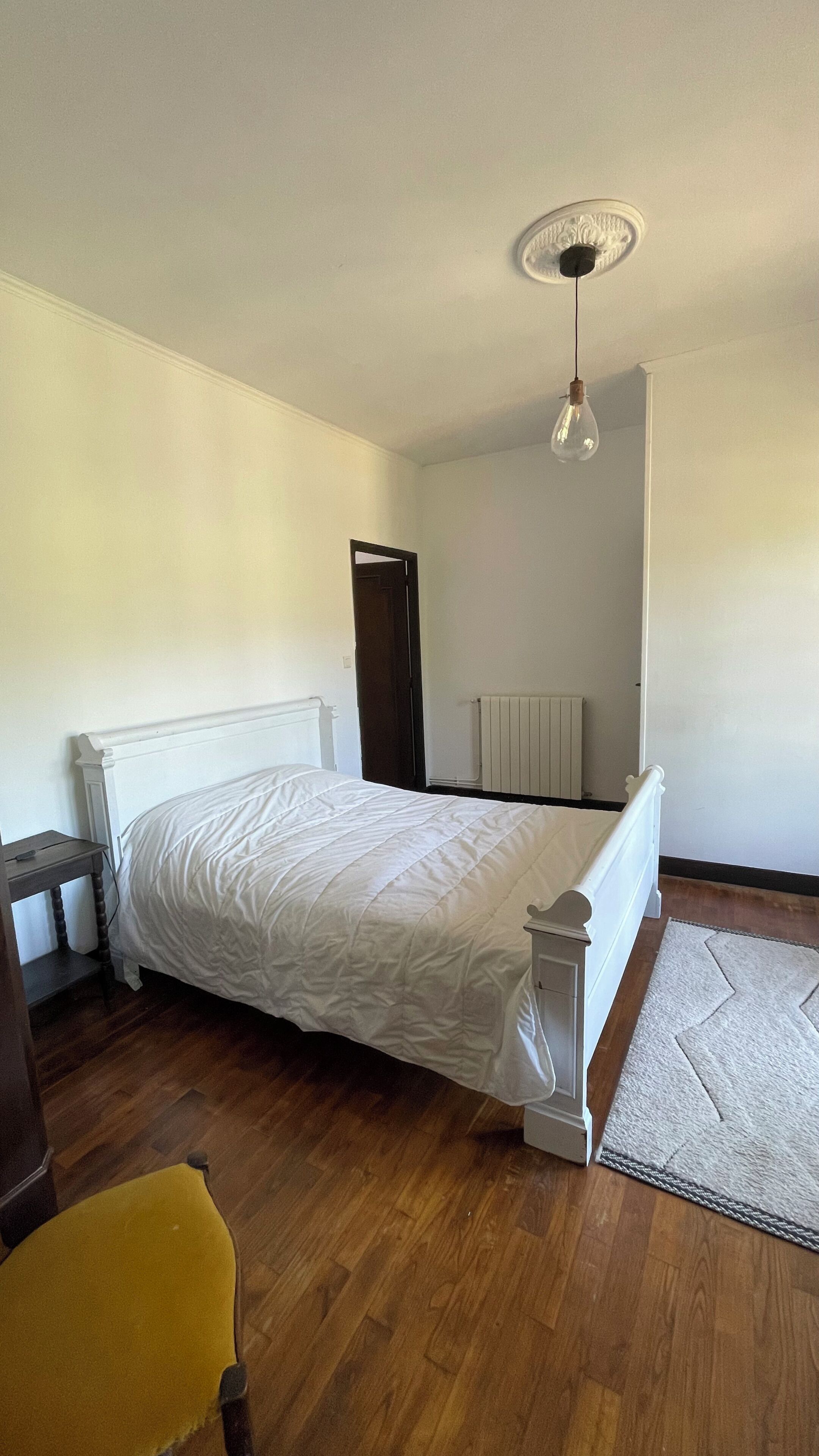 A bedroom with a bed, a wooden floor, a radiator, a door, and a hanging light fixture.