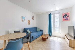 Ein modernes Wohnzimmer mit einem blauen Sofa, einem Holztisch, einem Fernseher auf einem Holzständer und einem Esstisch mit Stühlen.