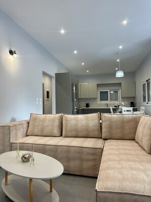 Ein modernes Wohnzimmer mit einem beigefarbenen Ecksofa, einem runden weißen Couchtisch, Deckenleuchten und Blick in die angrenzende Küche.