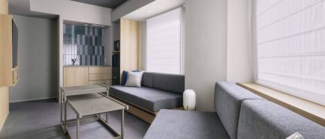 A modern, compact living space with a grey sofa, a small table, and a kitchenette area.