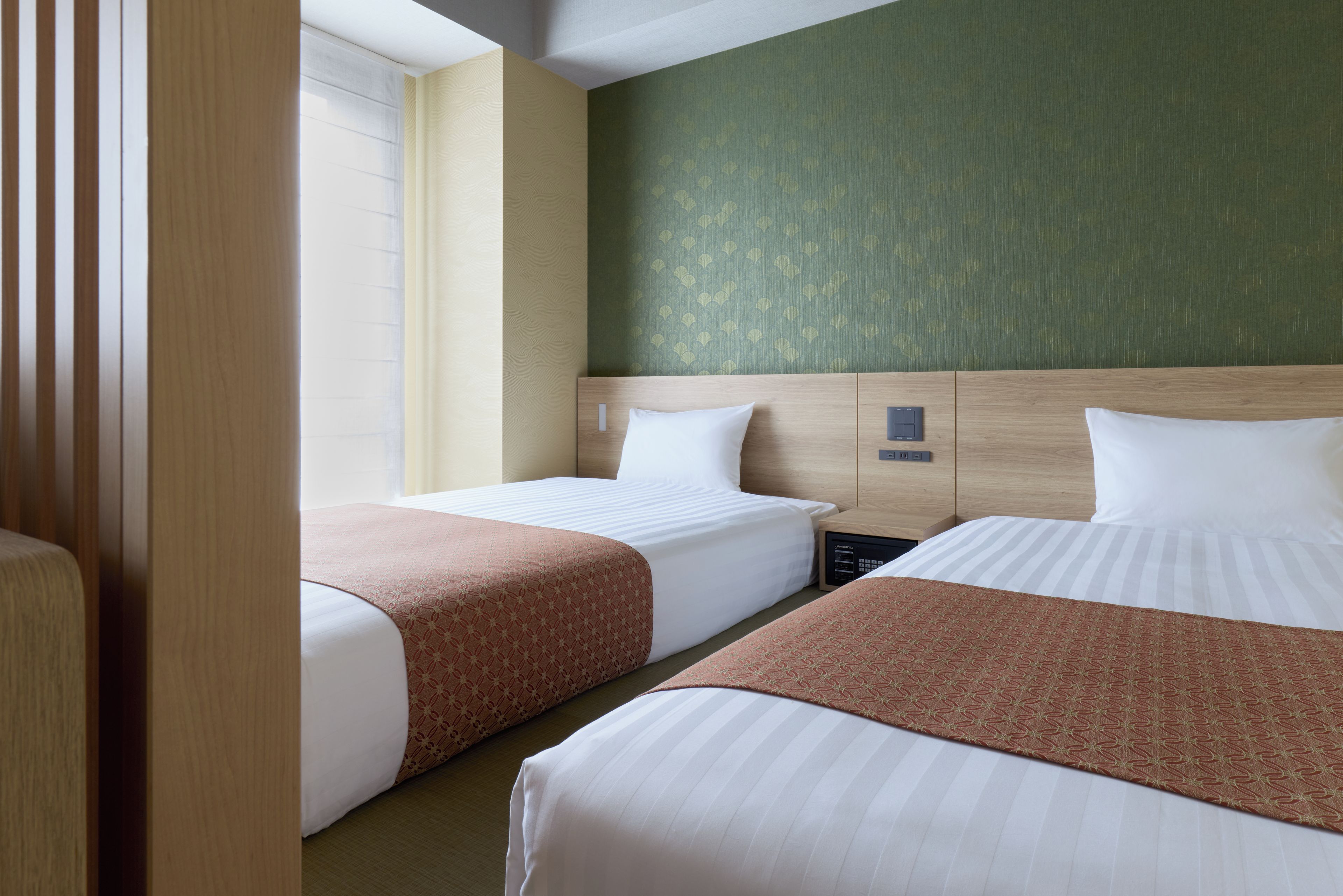 A hotel room with two beds, a nightstand, a control panel, and a window with blinds.