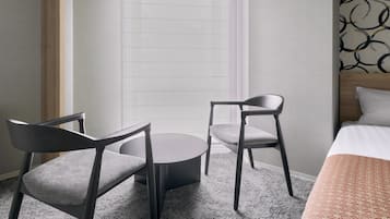 A hotel room with a bed, two chairs, a round table, and a window with sheer curtains.