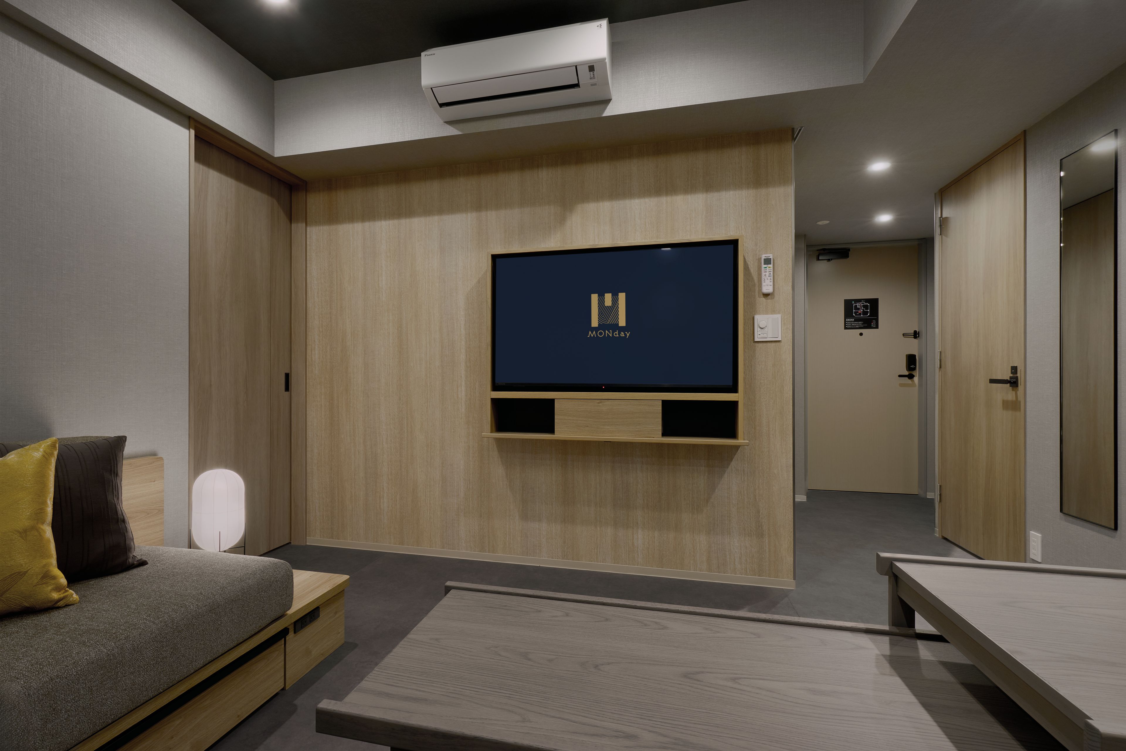 Ein modernes Hotelzimmer mit einem Wandfernseher, einer Couch mit Kissen, einem Nachttisch und einer Klimaanlage.