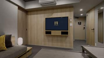 Ein modernes Hotelzimmer mit einem Wandfernseher, einer Couch mit Kissen, einem Nachttisch und einer Klimaanlage.
