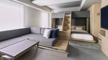 Una habitación moderna y funcional con un sofá gris, una mesita pequeña, una litera con escalera de madera y un televisor fijado a la pared.