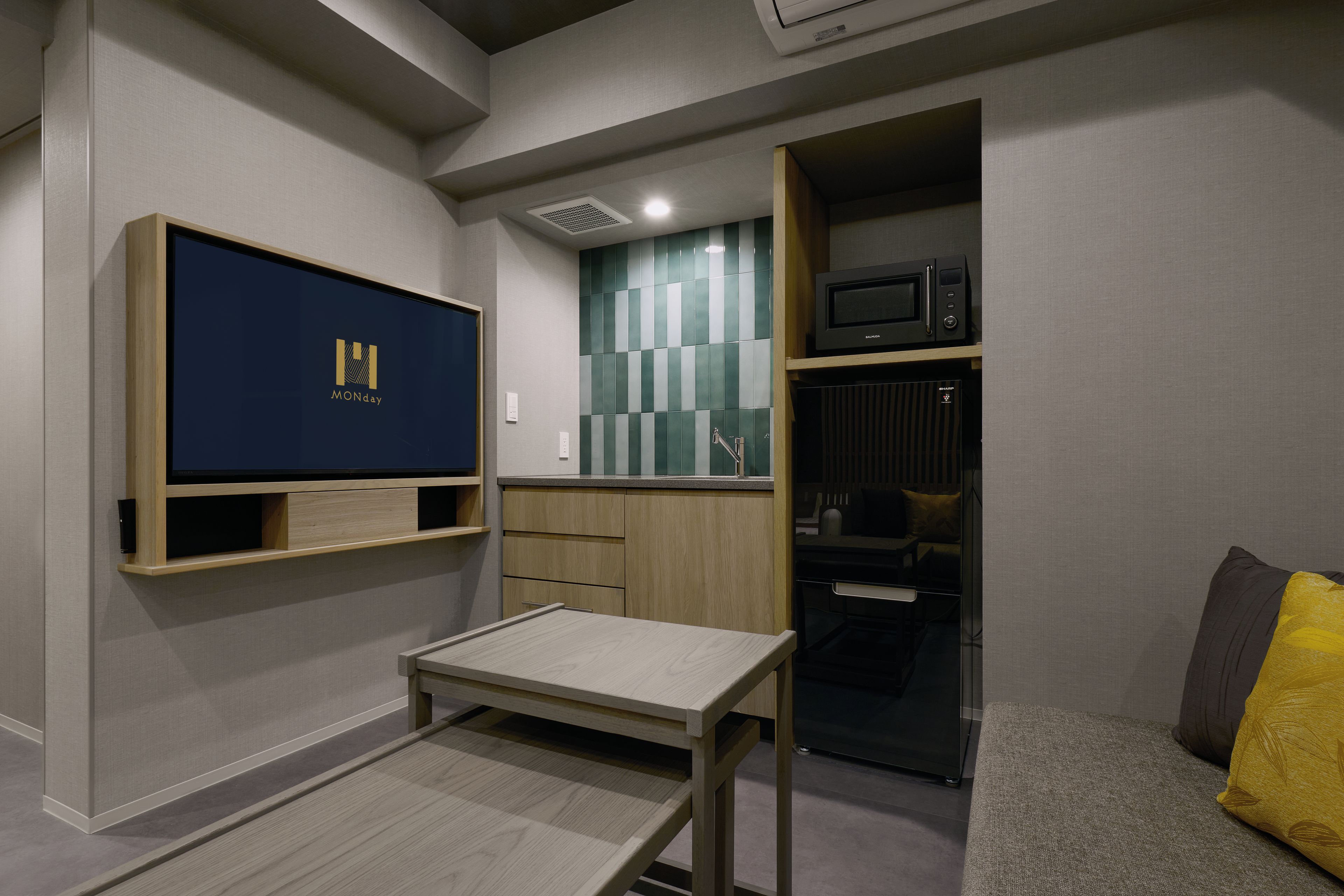 A compact living space with a flat-screen TV mounted on the wall, a small wooden table, a microwave, and a kitchenette area.