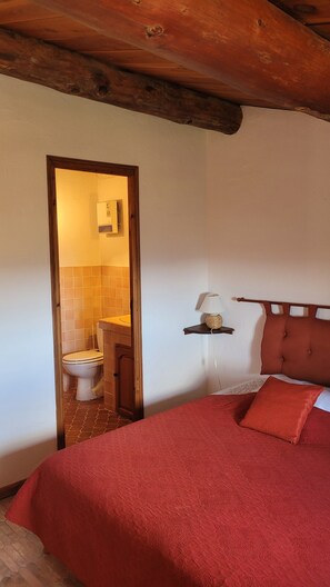 Ein Schlafzimmer mit Holzboden, einer roten Bettdecke, einer Nachttischlampe und Blick ins Badezimmer.