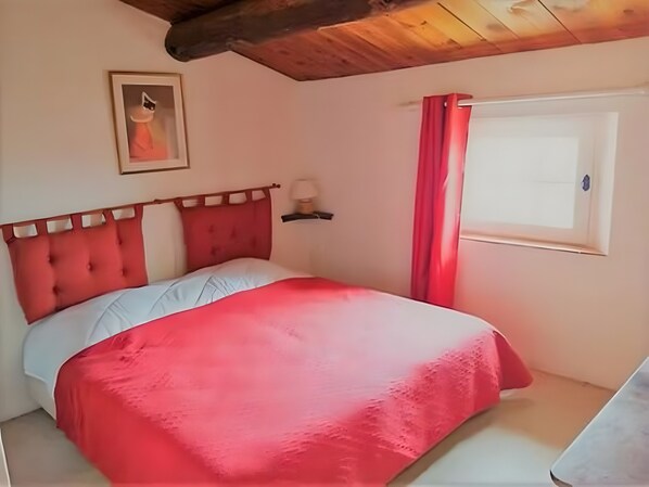 Ein Schlafzimmer mit rotem Bettbezug, einem Holz-Kopfteil, einem Fenster mit roten Vorhängen und einem gerahmten Bild an der Wand.