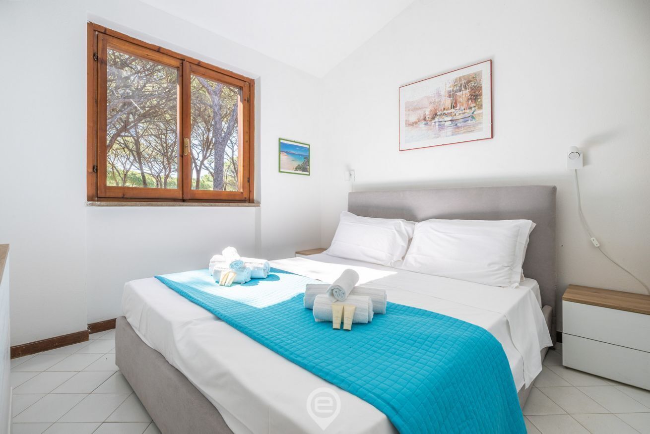 A bedroom with a bed, a window with a view of trees, a bedside table, and a wall-mounted lamp.