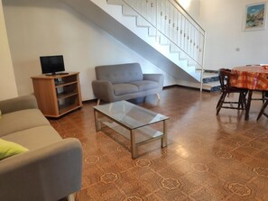 A living room with a sofa, a glass coffee table, a TV on a wooden stand, a staircase, and a dining table with chairs. - Beachside family villa with large, enclosed garden (Lignano Sabbiadoro)