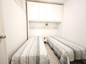 Ein kleiner, schmales Zimmer mit zwei Einzelbetten, einem Nachttisch, einer Lampe und einem weißen Kleiderschrank.