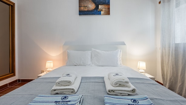 Et hotelværelse med en seng, to puder, to sengeborde med lamper, et billede på væggen og håndklæder med hotellets logo.