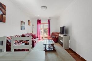 Ein Wohnzimmer mit einer Couch, einem Couchtisch, einem Fernseher und einem Balkon mit roten Vorhängen.