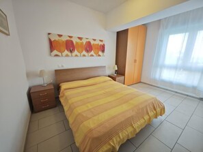 Un dormitorio con cama, mesita de noche, armario, un cuadro en la pared y una ventana con cortinas.