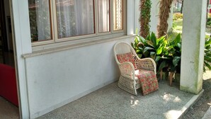 A room with a large window, a wicker chair with a floral cushion, and a concrete pillar. - Ideal property for group getaways in Lignano (Lignano Sabbiadoro)