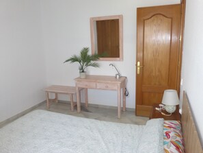 Ein Schlafzimmer mit einem Bett, einem Nachttisch mit Lampe, einem Holztisch mit Spiegel, einer kleinen Bank und einer Topfpflanze.