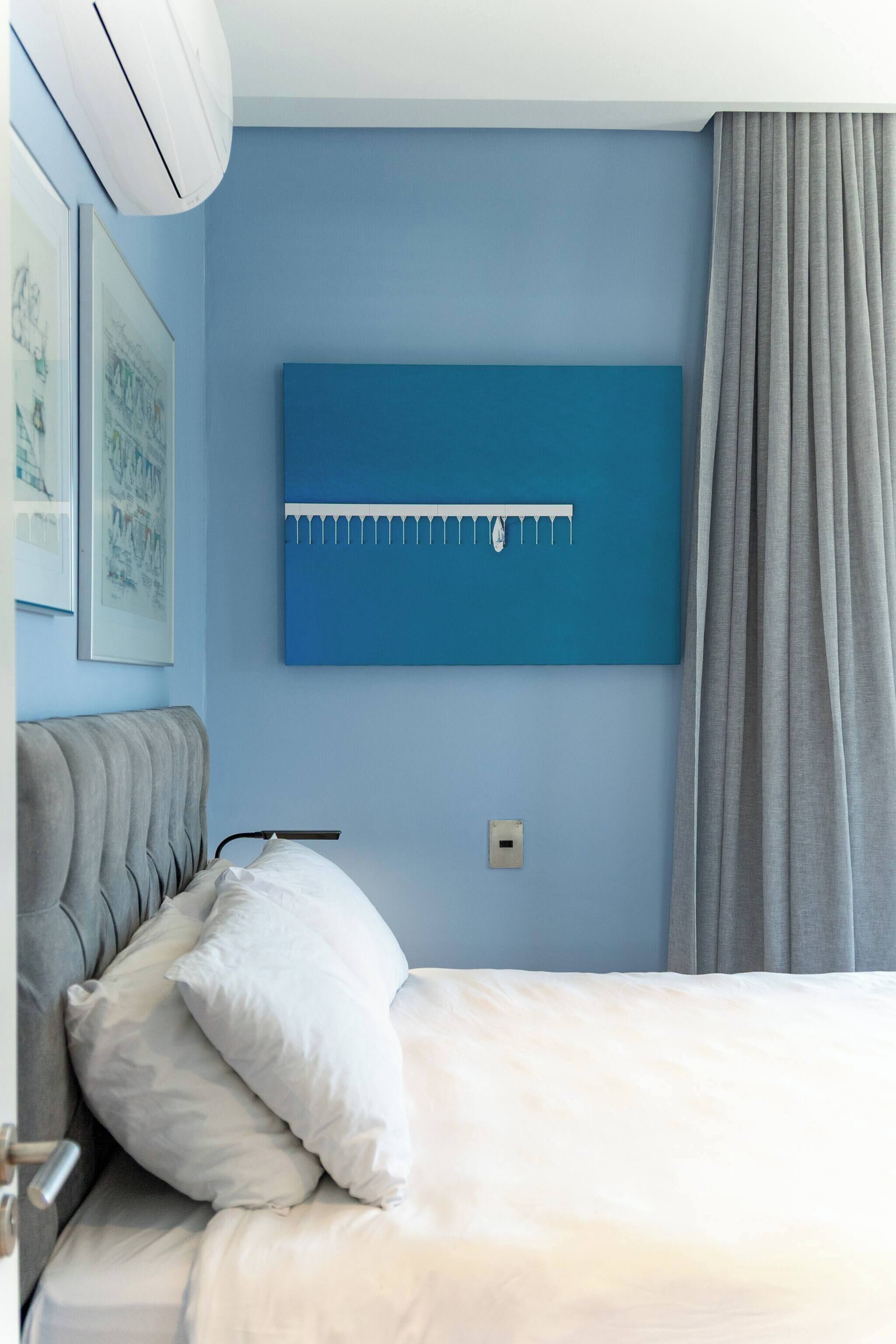 A bedroom with a bed, a wall-mounted air conditioner, a blue wall art piece, and a gray curtain.
