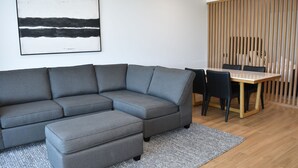 A modern living room with a grey sectional sofa, a small ottoman, a wooden dining table with chairs, and a vertical wooden partition. - Kove Apartment - G05 (Kiama)
