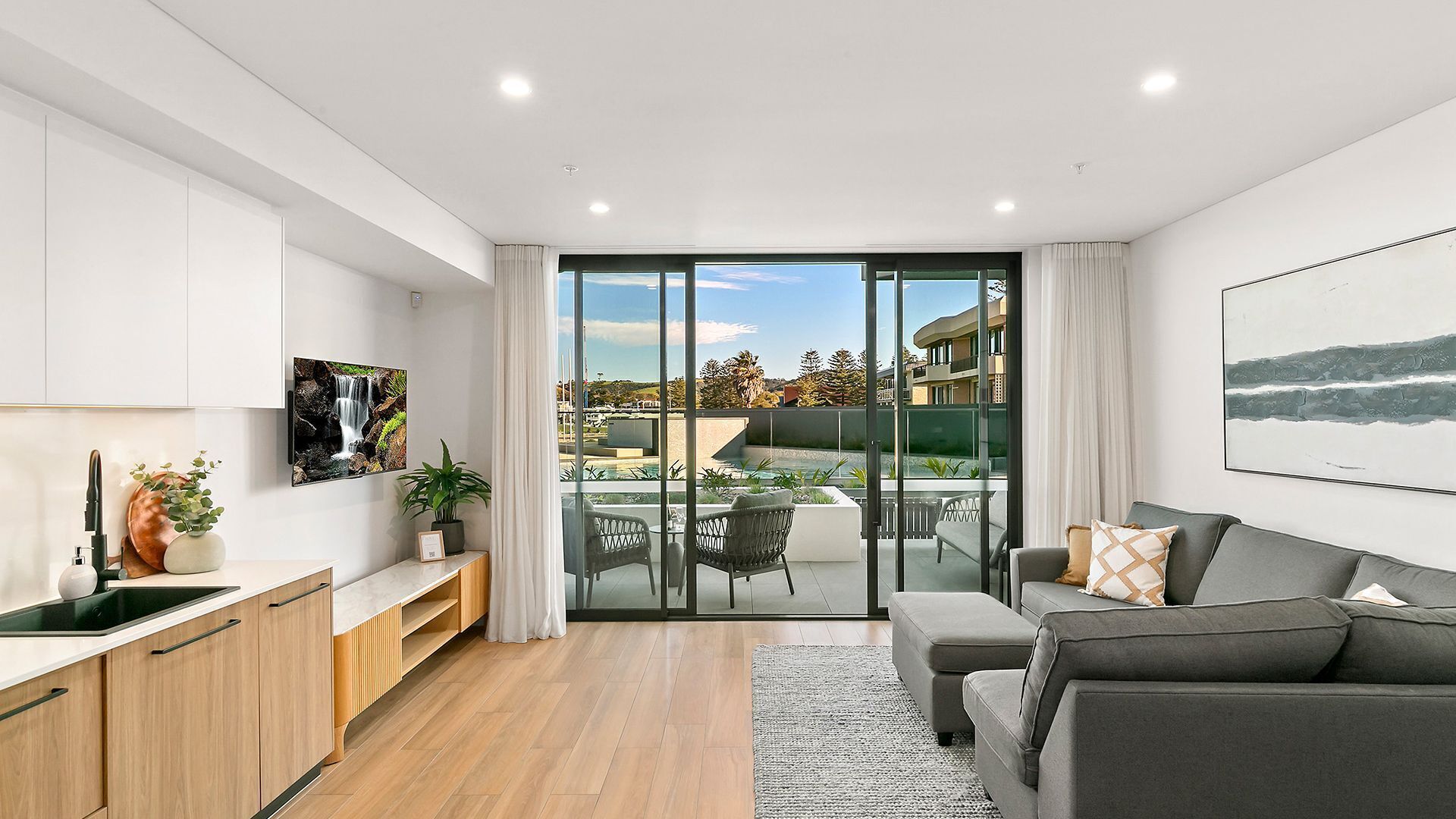 A modern living room with a sofa, a dining area with a table and chairs, a kitchen with cabinets and a sink, and a large sliding glass door leading to an outdoor patio with a pool and a view of buildings.