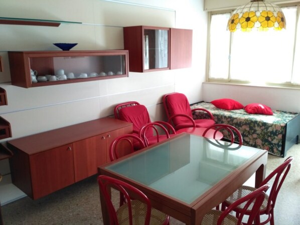 Une pièce de taille modeste, avec une table à plateau en verre, des chaises rouges, un canapé avec des coussins rouges, un buffet contenant de la vaisselle et un luminaire suspendu.