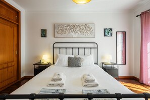 Un lit bien fait, avec deux serviettes, une tête de lit en métal, des tables de chevet avec des lampes et des œuvres d’art accrochées aux murs.