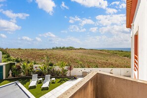 A balcony with a view of a field and ocean, white lounge chairs, and a small garden area.