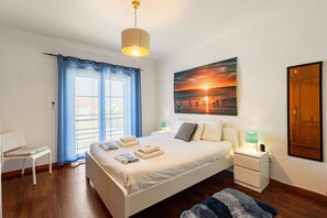 A bedroom with a bed, a nightstand, a chair, a window with curtains, a painting, and a wooden door.
