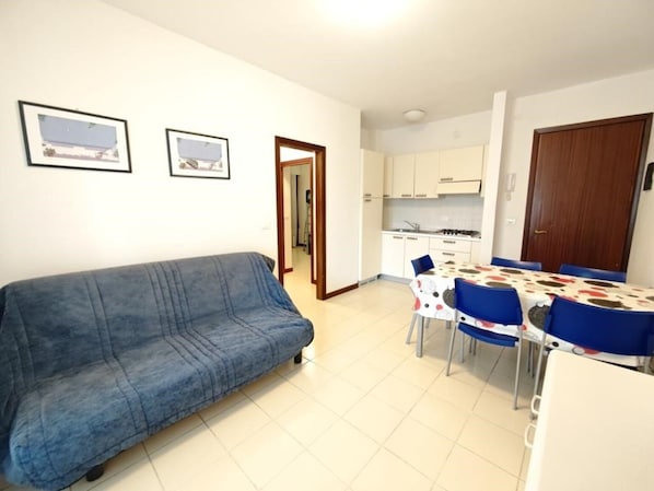 Ein Wohnzimmer mit Sofa, ein Essbereich mit Tisch und Stühlen, eine Küche mit Schränken und Herd, und eine Tür zu einem weiteren Raum.