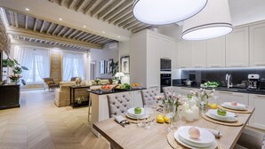 Una cocina moderna con armarios blancos, un comedor con mesa de madera dispuesta para una comida, y una sala de estar con sofá y cortinas.