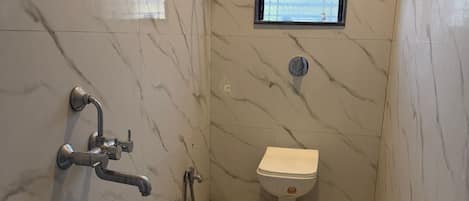 A bathroom with marble walls, a toilet, a shower with a hose, a bucket, and a window.